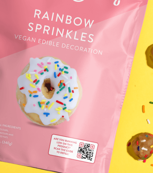 Eine rosafarbene und gelbe Streusel-Verpackung mit dem Bild eines weißen glasierten Donuts mit Streuseln und einem QR Code.